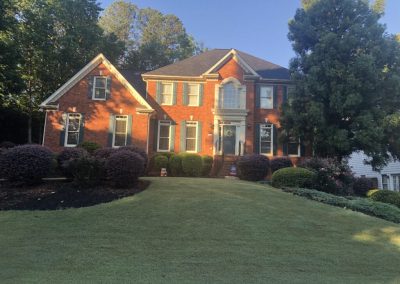 A brick house surrounded by a large yard, flower beds, and trees.