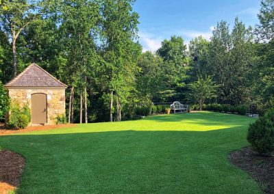 An small brick house surrounded by a huge yard full of trees and flower beds.