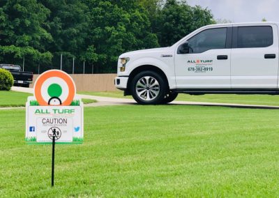 A white All Turf truck parked in a grassy area next to a pesticide application caution sign.