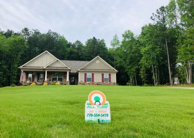 An All Turf sign in the middle of a yard in front of a house that is surrounded by trees.