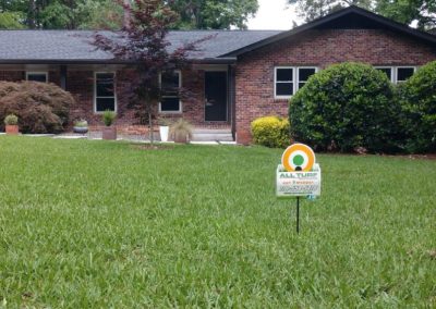 An All Turf sign in the middle of a yard in front of a red brick house.