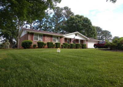 An All Turf sign in the middle of a flawlessly maintained yard in front of a house surrounded by trees.