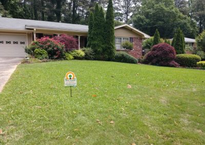 An All Turf sign in the middle of a yard in front of a house that is surrounded by trees.