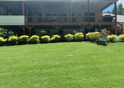 A two-level wooden house surrounded by bushes and green areas.